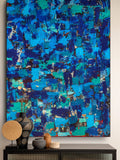 A large abstract canvas painting with a textured, layered brushstroke pattern in vibrant blues, teals, and deep navy tones, displayed on a wall above a black console table with decorative vases.
