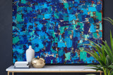 Large abstract wall art featuring a textured mosaic of blue, teal, and navy brushstrokes with gold leaf accents, displayed above a console table with decorative vases and a green plant.