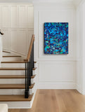 An abstract canvas painting featuring vibrant shades of blue, teal, and gold, displayed on a white paneled wall in a modern staircase hallway with wooden steps.